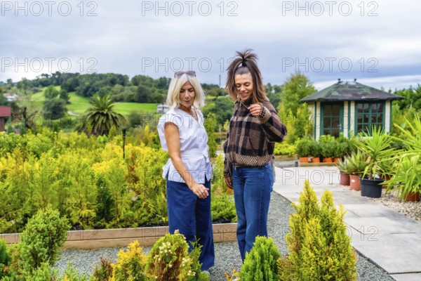 Mother and daughter browsing potted plants and small trees at a garden center, enjoying a relaxed day together selecting greenery and sharing a warm, casual moment outdoors