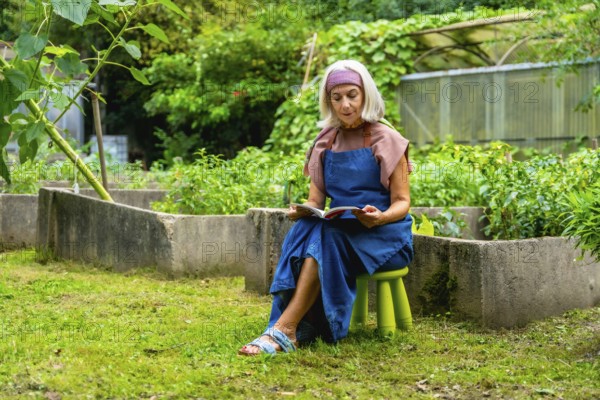 Senior woman wearing an apron and headband, enjoying a quiet moment reading a book while sitting on a stool in her green outdoor garden next to raised beds