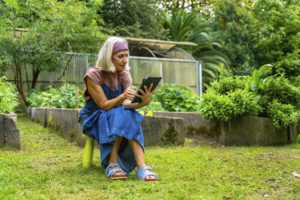 Senior woman sitting on a stool in her green garden, using a digital tablet for managing her plants and relaxing outdoors in nature with modern technology