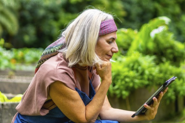 Senior woman with blond hair and a pink headband enjoying leisure time in a lush green garden, comfortably reading or browsing content on a digital tablet