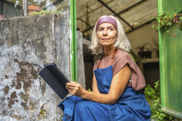 Senior woman in apron sitting by a greenhouse entrance, holding a digital tablet and looking thoughtfully, managing her small business and plant inventory