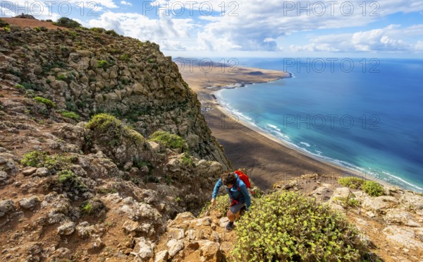 Female hiker climbing to Castillejo summit, view from the Risco de Famara cliffs of the coast and the sea with Famara beach, Playa de Famara with La Calaeta, Lanzarote, Canary Islands, Spain