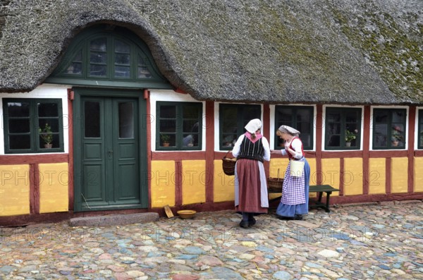 Two women in older farm costumes in front of a half-timbered house with a thatched roof and on paved ground in Den gamle by, The old village, open air museum in Odense, Fyn island, Fuen, Denmark, Scandinavia