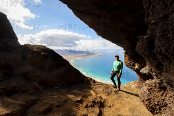 Tourist in the Cueva de las Cabras cave, young woman enjoying the view from the Risco de Famara cliff to Famara beach, Playa de Famara with La Calaeta, Lanzarote, Canary Islands, Spain