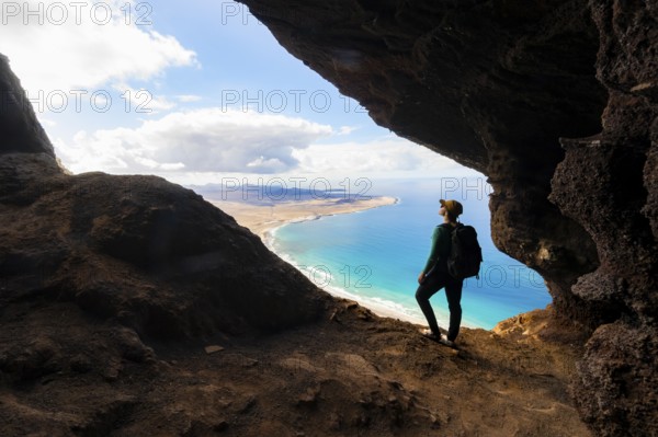 Tourist in the Cueva de las Cabras cave, young woman enjoying the view from the Risco de Famara cliffs on Famara beach, Playa de Famara with La Calaeta, Lanzarote, Canary Islands, Spain