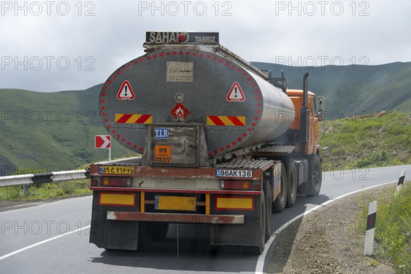 A tank truck drives on a winding road through a mountainous and green landscape under cloudy sky, truck with Iranian license plate, Syunik province, Armenia