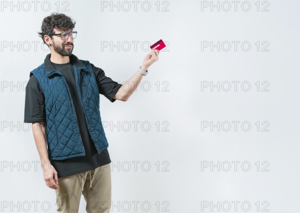 Unsure and confused young man holding and looking at a credit card. Confused modern guy holding a credit card isolated