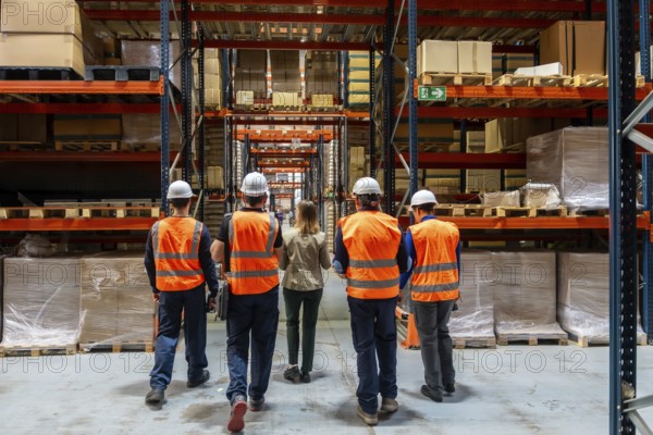 Warehouse manager and four workers wearing safety vests and hard hats walking through an industrial storage facility, examining rows of shelves filled with goods and packages