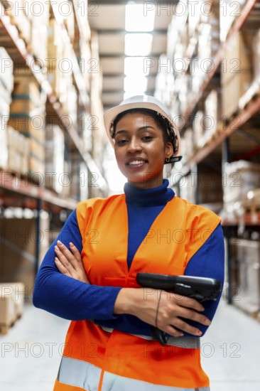 Confident black woman worker wearing a hardhat and high visibility vest, holding a barcode scanner in a modern logistics and distribution warehouse environment with stacked boxes