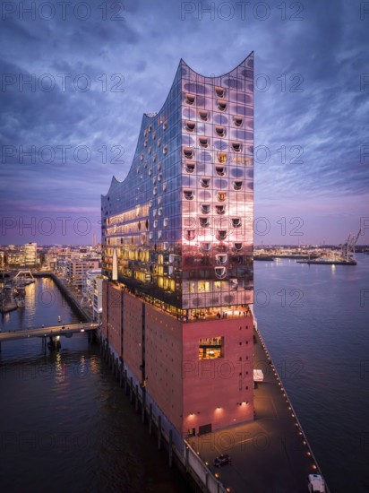 Aerial view of the Elbe Philharmonic Hall with reflections on the façade in evening light above the harbor, surrounded by water and urban backdrop, Hamburg, Germany