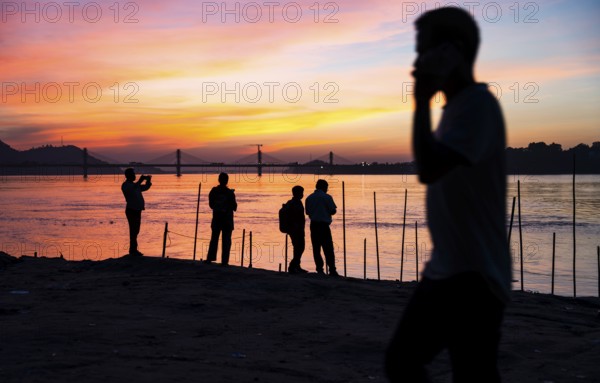 People enjoy the vibrant sunset by the Brahmaputra riverside, capturing photos and moments against the scenic backdrop of a bridge and glowing sky, on November 4, 2025 in Guwahati, India