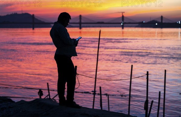 A person browsing his mobile phone by the Brahmaputra riverside, during sunset, on November 4, 2025 in Guwahati, India