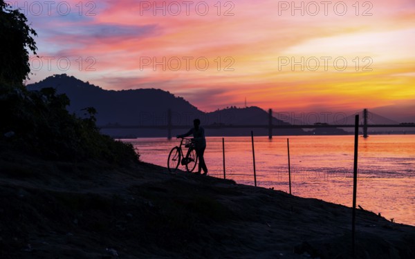 A man pushes his bicycle along the Brahmaputra riverbank during a vibrant sunset, with the silhouette of a bridge and hills adding to the serene evening atmosphere., on November 4, 2025 in Guwahati, India