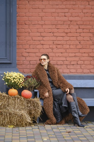 A woman sits on a bench dressed in warm fur coat. She poses near colorful pumpkins and vibrant flowers against a red brick wall, embodying the autumn spirit