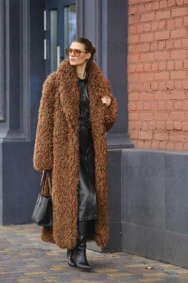 A fashionable woman strolls along a city sidewalk wearing a warm brown fur coat and stylish black attire. She holds a handbag and showcases confidence near a brick building in the background
