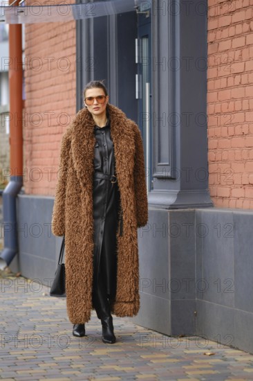 A fashionable woman strolls along a city sidewalk, wearing a cozy brown fur coat. She pairs it with a chic black outfit, sunglasses, and confidently walks past a striking red brick building