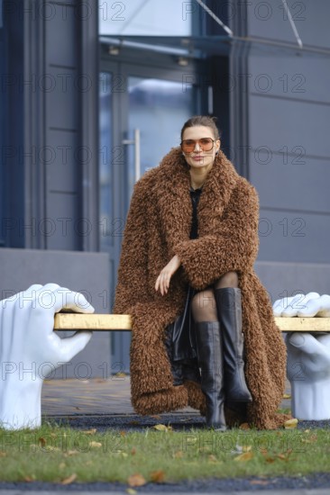 A stylish woman wearing a brown fur coat and black knee-high boots poses on a unique bronze bench supported by large sculpted hands. The background features a modern building