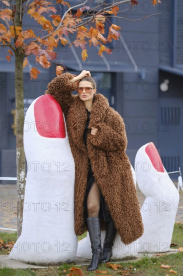 A fashionable woman in a cozy brown coat stands near large white sculptures resembling fingers with red nails. Colorful autumn leaves surround her as she strikes a confident pose outdoors
