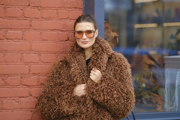 A woman poses confidently in a warm, brown, fluffy coat and oversized sunglasses. She stands against a red brick wall, showcasing a modern urban style in a vibrant setting