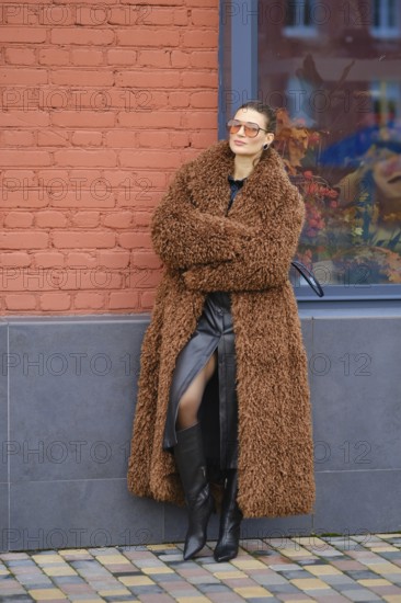 A fashion model stands with crossed arms beside a red brick wall. She wears a fluffy brown coat, a black dress, and knee-high black boots, appearing stylish and relaxed in a vibrant urban setting