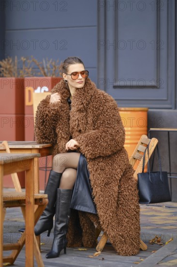 A stylish woman is seated at a wooden table outside a cafe, wearing a fluffy brown coat and oversized sunglasses. She turns her head to the side and looks into the distance, waiting for someone