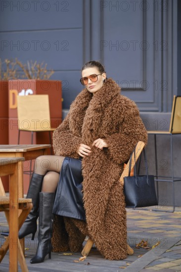 A stylish woman sits at an outdoor cafe during autumn, wearing a fuzzy brown coat and black knee-high boots. She pairs her look with oversized sunglasses and handbag