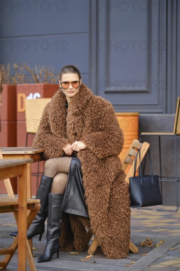 A woman sits comfortably at a wooden cafe table, wearing a brown fluffy coat and knee-high black boots. She enjoys the warm sunny autumn weather