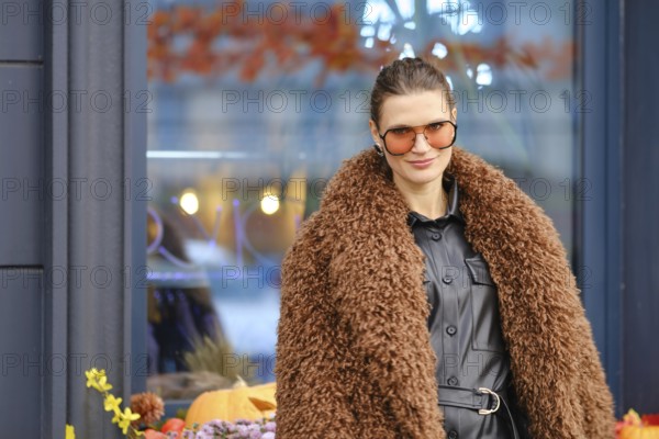 A woman wearing a brown furry coat and sunglasses stands outside a cozy cafe filled with autumn decorations. Her confident smile reflects the vibrant atmosphere of the season