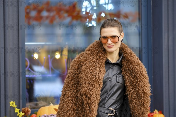 Fashionable woman in stylish brown fur coat posing outside a modern cafe on a sunny autumn day