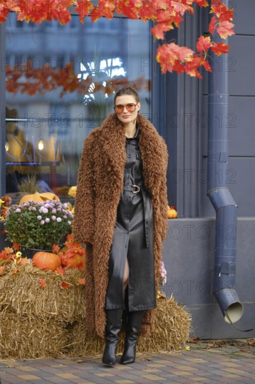 A woman dressed in a stylish brown fuzzy coat and black leather dress stands next to a seasonal display of pumpkins and autumn leaves