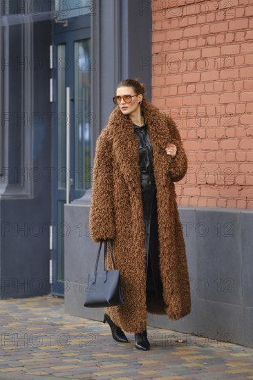 A stylish woman with sunglasses walks along a sidewalk by a vibrant brick wall. She wears a fluffy brown coat over a sleek outfit and carries a black handbag while enjoying the day