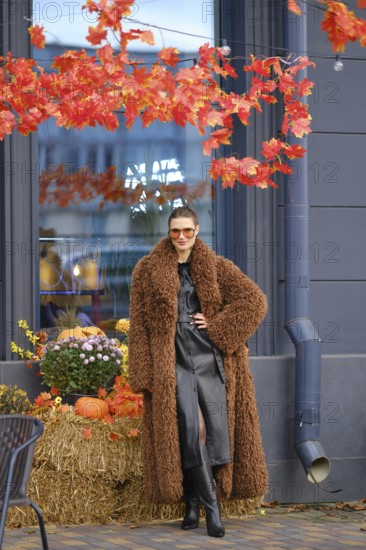 A stylish model stands near a store with bright autumn leaves overhead. She wears a brown fur coat and sunglasses. Pumpkins and flowers add seasonal charm to the cozy setting