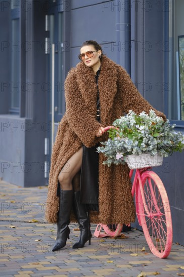 A fashionable woman stands against a modern building, wearing a cozy brown coat and tall black boots. She leans on a pink decorative bicycle adorned with fresh flowers, enjoying her city surroundings