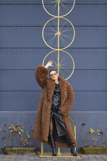 A woman stands confidently near a striking bike wheel art piece. She wears a fluffy brown coat and sleek black outfit