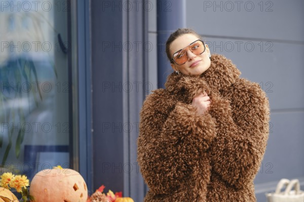A young woman stands outside a shop, dressed in a fluffy brown coat and stylish sunglasses. Colorful pumpkins decorate the scene, capturing the essence of autumn in a cheerful atmosphere