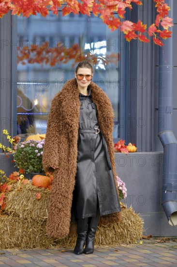 A stylish woman poses outside a farmer's grocery shop decorated for autumn, wearing a brown fur coat and sunglasses. Colorful pumpkins and flowers add to the seasonal charm