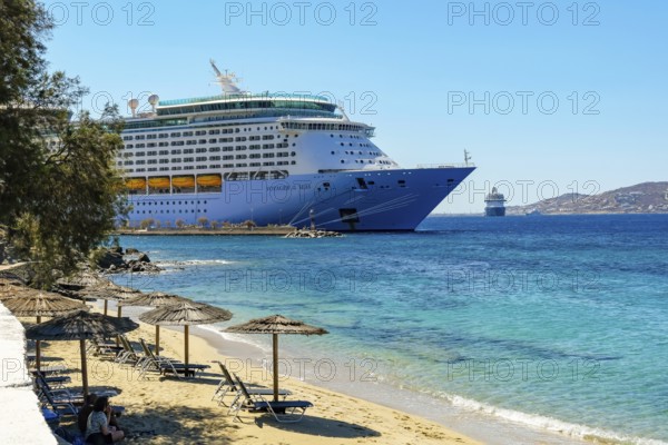 Mykonos, Cyclades, Greece - Voyager of the Seas cruise ship is located in Mykonos Harbour Bay. Front beach chairs and umbrellas on the sandy beach of Paralia Agios Stefanos