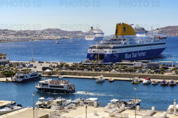 Mykonos, Cyclades, Greece - Mykonos New Port, a Blue Star Ferries ferry, is located on the quay in the new port in front of the skyline of Mykonos Town. In front, a taxi boat a sea bus takes tourists to the old town. At the back, there are more cruise ships in the bay in front of Mykonos City with a view of the old town with the windmills. Cruise ships bring tourists to the old town
