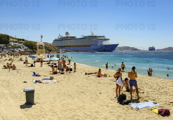Mykonos, Cyclades, Greece - Voyager of the Seas cruise ship is located in Mykonos Harbour Bay. In front, tourists and locals bathe on the sandy beach of Paralia Agios Stefanos