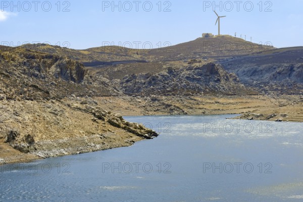 Mykonos, Cyclades, Greece - Dry Fokos Reservoir near Ano Mera. The Mykonos Municipal Water Supply and Sewage Management Company has two dams, Marathi Reservoir and Fokos Reservoir, to supply water to the island. Due to the prolonged drought, both are almost empty and are not being used. A wind turbine at the back of the mountain