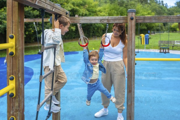 Two brothers and their mother are having fun on a playground jungle gym, with one child climbing a rope ladder and the other hanging from rings