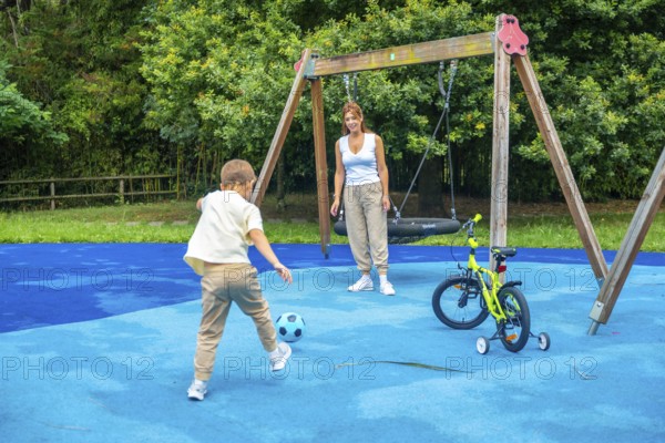 Young boy kicking a soccer ball on a playground while mother watches, with a swing set and bicycle nearby, creating a joyful scene of family fun on a sunny day