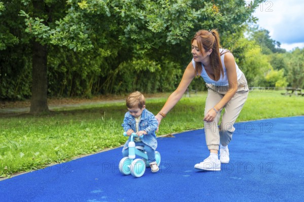 Young mother helping her son learn to ride a balance bike on a blue surface in a green park, promoting childhood development and family bonding