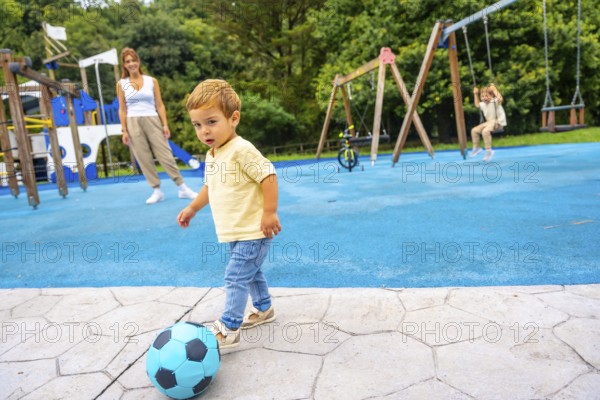 Happy toddler kicking a vibrant soccer ball at the playground, with mother and sister enjoying playtime in the background, creating a joyful scene of family togetherness and fun