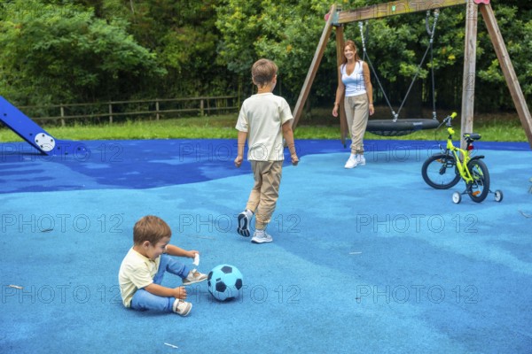 Two brothers are playing with a ball and a bicycle at the playground while their mother watches them, promoting family bonding and outdoor fun