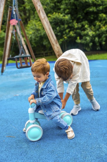 Older brother is helping his younger brother to ride a balance bike on a blue playground surface with a swing set in the background, promoting childhood fun, learning, and brotherly bonding