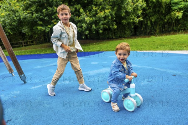Two brothers are spending time together on a playground, one riding a balance bike and the other walking next to him on a blue rubber surface