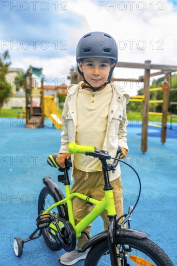 Happy boy wearing helmet learning to ride a bicycle with training wheels in a playground on a sunny day, promoting children's road safety and outdoor activities