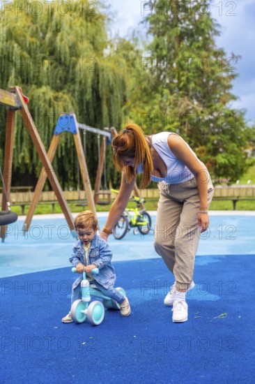 Young mother helping her toddler son learn to ride a balance bike on a playground's soft blue surface, promoting early childhood development and family bonding