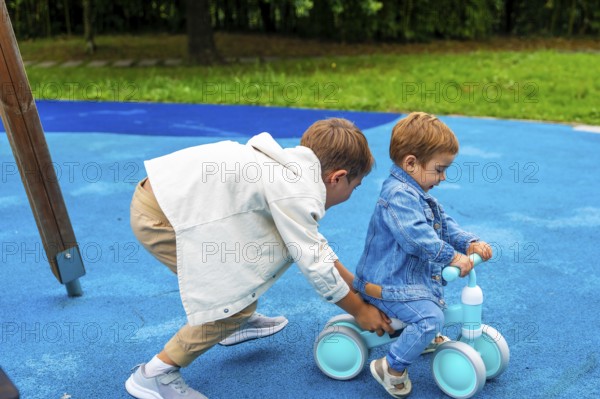 In a vibrant playground setting, an older brother supports his younger sibling as he learns to ride a balance bike, fostering a sense of camaraderie and shared joy in outdoor play